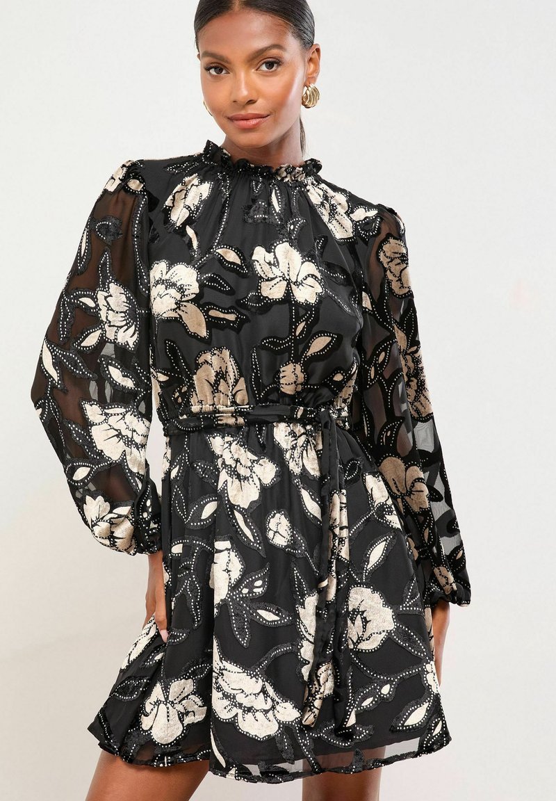 Rochie neagră cu flori, cu mâneci transparente, decolteu volănaș și talie evidențiată cu brâu. Prezintă modele florale metalice și accente texturate.