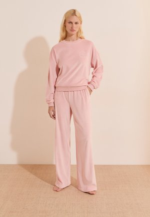 Donna in piedi a piedi nudi che indossa una felpa rosa a maniche lunghe larghe e pantaloni abbinati a gamba larga davanti a una parete chiara e liscia.