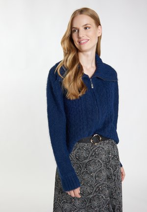 Vrouw met lang golvend haar, gekleed in een marineblauw vest met ritssluiting en een patroonrok met een zwarte riem, tegen een eenvoudige lichte achtergrond.