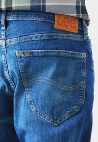 Pantalones vaqueros de mezclilla azul con un corte clásico, que cuentan con una etiqueta de cuero con el logotipo de Lee, costuras en contraste y un diseño de bolsillo trasero curvado.