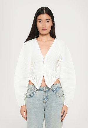 White cropped cardigan with a V-neck, featuring textured pleats, front buttons, and puffed long sleeves, worn over light blue jeans.