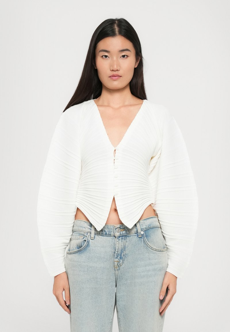 Witte cropped cardigan met een V-hals, voorzien van gestructureerde plooien, knopen aan de voorkant en eengeflonken lange mouwen, gedragen over lichtblauwe jeans.