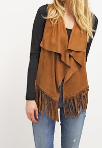 Chaleco de ante marrón con parte delantera drapeada, con detalles de flecos en el dobladillo; diseño en capas usado sobre una blusa negra de manga larga y jeans azules.