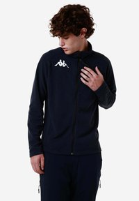 Giacca in pile blu navy con colletto alto e zip completa. Presenta un logo bianco sul lato sinistro del petto e un tessuto testurizzato con una finitura liscia.