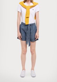 Person som bär en vit pikétröja med en gul tröja över axlarna, blå shorts med bälte och vita sneakers som står framför en vit bakgrund.