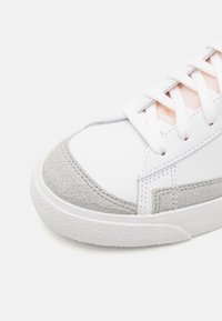 Vit sneaker med en slät läderövre, grå mocka på tårna, vita snörning och en texturerad gummisula. Minimalistisk design med pastellaccenter.