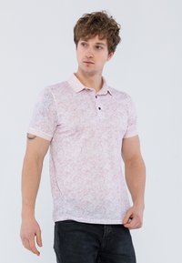 Světle růžové polo tričko s texturovaným vzorem, krátkými rukávy a límečkem. Obsahuje tři černé knoflíky podél zapínání.