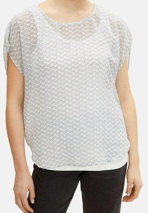 Kobieta ubrana w białą bluzkę z krótkim rękawem z czarnym wzorem w kształcie muszelek oraz czarne spodnie, stojąca na tle jednolitego tła.