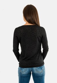 Donna con lunghi capelli marroni che indossa una camicia nera a maniche lunghe con stampa leopardata aderente e jeans blu, in piedi con le mani dietro la schiena.