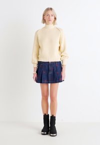 Pull en maille côtelée crème à col montant, associé à une jupe en denim foncé avec broderies florales rouges et des bottines noires avec des accents argentés.