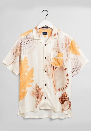 Mies, joka käyttää vaaleaa lyhythihaista paitaa, jossa on simpukkakuosia, sinapinkeltaisia shortsseja ja mustia sandaaleja, seisoo mauve-taustaa vasten.