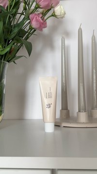 Tube of Korean skincare cream on a white surface, flanked by pink and white flowers in a vase and three tall beige candles in a holder.