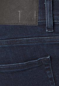 Jeans de mezclilla azul oscuro con un parche de cuero negro texturizado que presenta letras en relieve. Costuras visibles y un detalle en el bolsillo trasero.