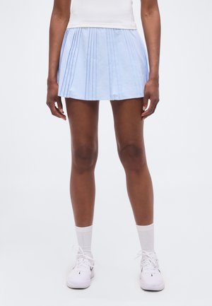 Personne portant une mini-jupe plissée bleu clair, un haut blanc, des chaussettes blanches montantes et des baskets blanches Nike, debout devant un fond uni.