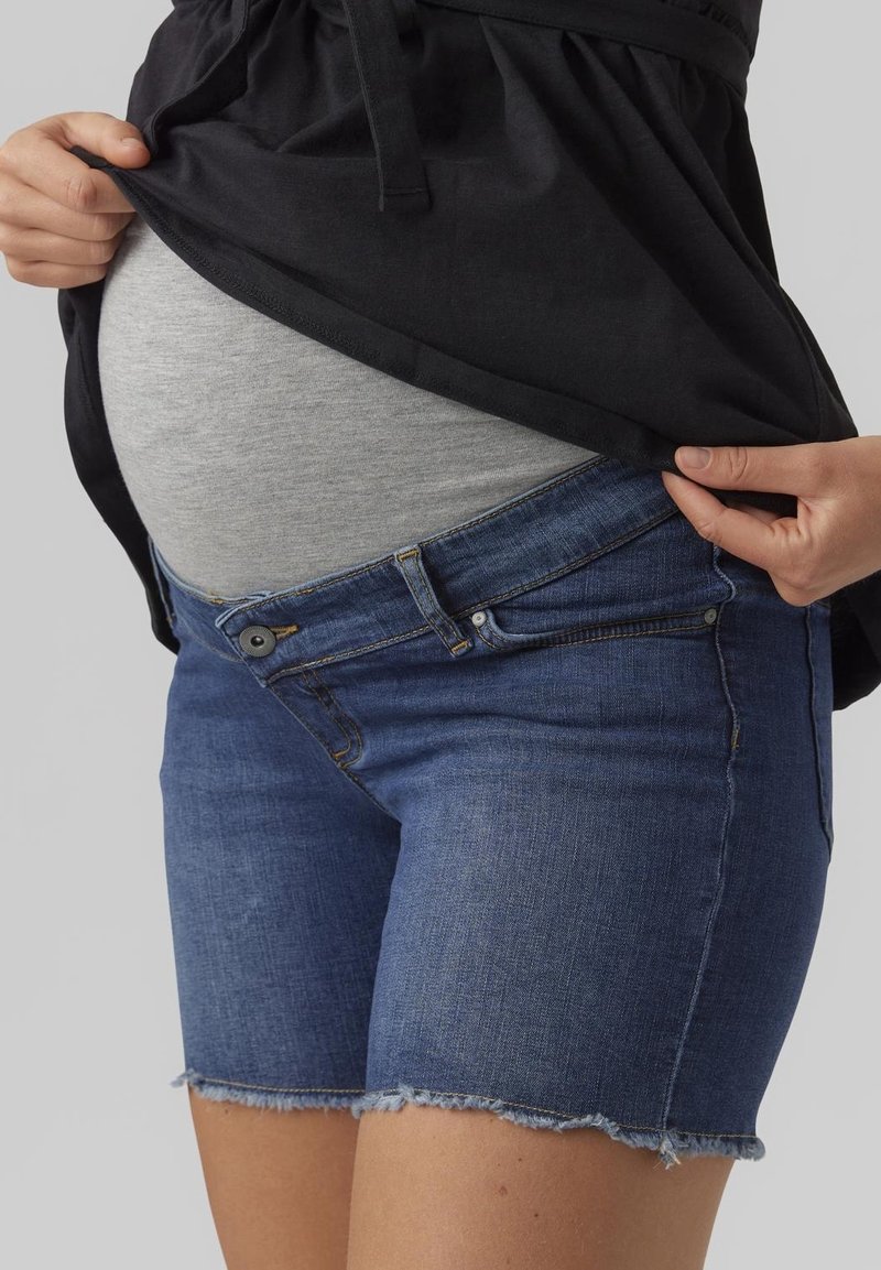 Jeans-Umstandsshorts mit tailliertem Bund, leichtem Used-Look und fransigem Saum. Kombiniert mit einem grauen Oberteil, das einen runden Bauch verdeckt.
