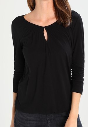 Femme portant un haut noir à manches longues avec une petite encolure goutte d'eau et des détails froncés au col.