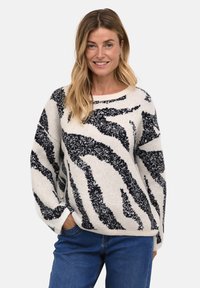 Pull surdimensionné en crème avec des rayures zèbre noires. Texture douce, poignets et ourlet côtelés. Associé à un jean bleu.