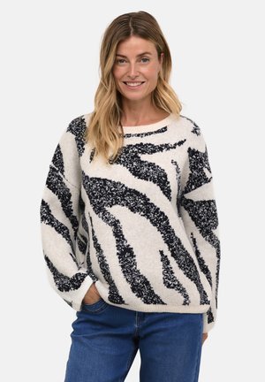 Pull surdimensionné en crème avec des rayures zèbre noires. Texture douce, poignets et ourlet côtelés. Associé à un jean bleu.