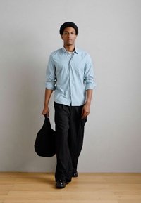 Ανοιχτό μπλε καρό πουκάμισο με κουμπιά, μαύρο παντελόνι με φαρδιές γραμμές, μαύρα παπούτσια, κρατώντας μια μαύρη τσάντα ώμου, στέκεται μπροστά από έναν απλό τοίχο.