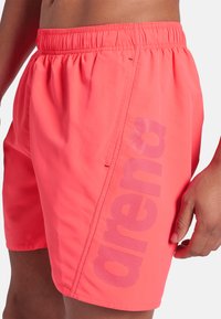 Primo piano di una persona che indossa dei pantaloni da bagno corallo brillanti con cintura elastica e il logo viola "arena" stampato verticalmente sul lato.