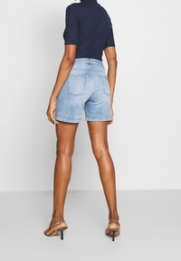 Mujer de espaldas vistiendo un short de mezclilla azul claro, una camiseta de manga corta color navy y sandalias de tacón de serpiente, sobre un fondo blanco plano.