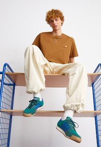 Brun bomull t-shirt med liten logotyp, krämfärgade baggy-byxor och grön-blå mockaskor med lila ränder. Modellen sitter på en trähylla.