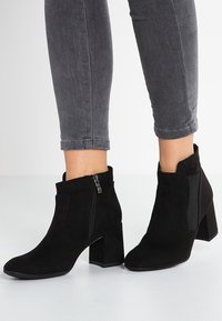 Botas de ante negras hasta el tobillo con tacones de bloque y cremalleras laterales, usadas con jeans ajustados gris oscuro.