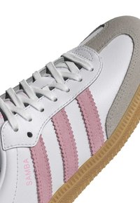 Vit sneaker med rosa sågade ränder, grå mockadetaljer, vita skosnören och gummisula. "SAMBA" tryckt på sidan.