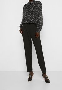 Zwarte blouse met een subtiele patroon ontwerp, gecombineerd met zwarte, high-waisted pantalon en zwart, puntige hakken met een gouden gespdetail.