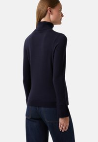 Frau in einem navyblauen Rollkragenpullover mit breiten Bündchen und dunkelblauen Jeans, von hinten vor einem schlichten Hintergrund gezeigt.