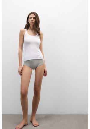 Junge Frau trägt ein weißes Tanktop und graue Unterwäsche und steht barfuß vor einer einfachen weißen Wand und einem grauen Boden.