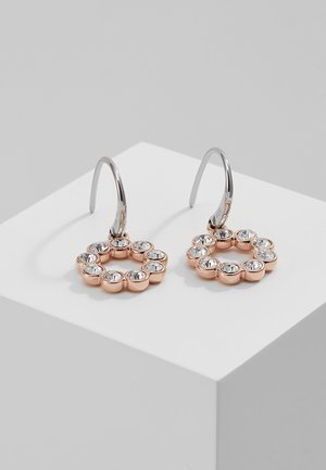 Boucles d'oreilles circulaires en or rose avec dix pierres précieuses transparentes chacune et crochets en argent sur un bloc géométrique blanc.