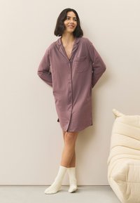 Camicia da notte oversize color malva con colletto, tasca frontale e bottoni. Indossata con calzini bianchi al ginocchio. Tessuto morbido e vestibilità comoda.