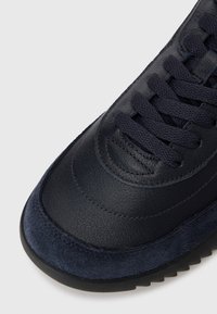 Sötétkék sneaker, bőrből és velúrból készült. Kerek orrú, egyszínű fűzővel rendelkezik, és a tapadást biztosító texturált talp jellemzi. Finom varrási részletek.