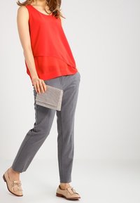 Donna che indossa un top senza maniche rosso, pantaloni grigi su misura, mocassini in pelle beige, mentre tiene una piccola borsa a tracolla con texture.
