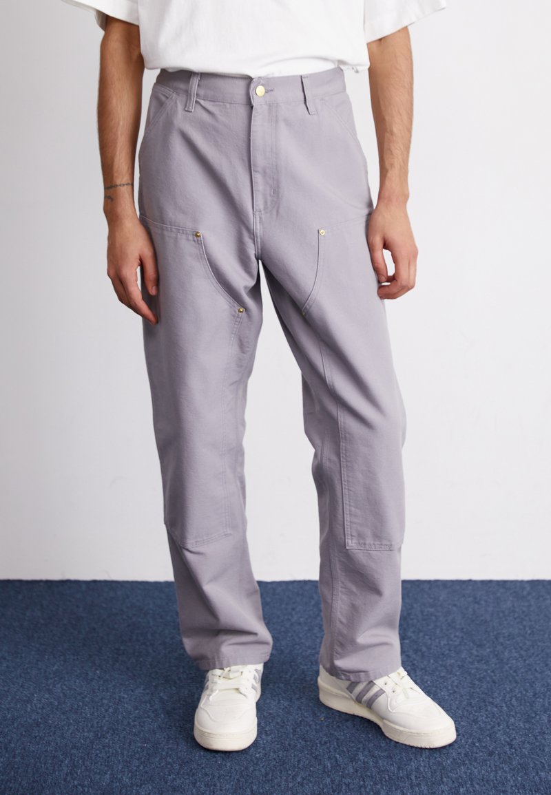 Hellgraue Baumwollhose mit lässiger Passform, ausgestattet mit goldenen Hardware-Details und mehreren Vordertaschen, kombiniert mit weißen Sneakers.