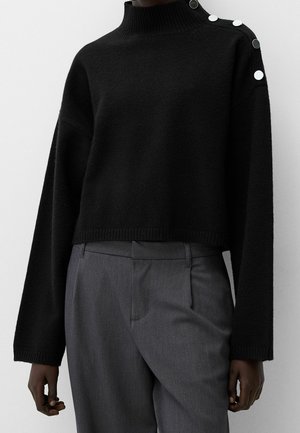 Pull court noir à col roulé avec des boutons blancs sur l'épaule gauche, associé à un pantalon gris taille haute et ajusté porté par une personne.
