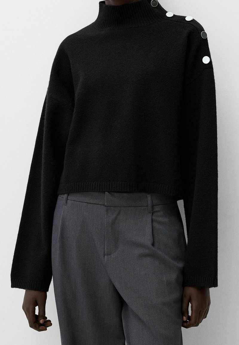 Pull court noir à col roulé avec des boutons blancs sur l'épaule gauche, associé à un pantalon gris taille haute et ajusté porté par une personne.
