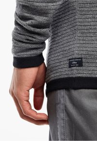 Grå texturerad tröja med ett randigt mönster, med svarta ärmslut och ett varumärkesmärke på sidan. Handen vilar på grå byxor.