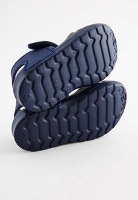 Scarpe per bambini di colore blu scuro con suola in gomma testurizzata dotata di un motivo a zigzag, cinturino in velcro e dettaglio del logo sul lato.