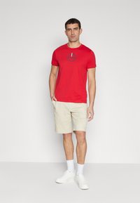 Rød t-shirt med blåt logo på brystet, parret med beige shorts og hvide sneakers. Modellen står foran en ensfarvet baggrund.
