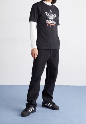 Black graphic t-shirt with white Adidas logo, layered over a white long-sleeve shirt. Paired with black jeans and black sneakers featuring white stripes.