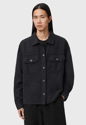 Jong persoon met lang zwart haar draagt een zwarte jack met knopen en twee borstzakken, staand tegen een eenvoudige lichte achtergrond.