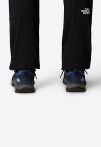 Siniset vaelluskengät tummanharmailla pohjilla ja keltaisilla yksityiskohdilla, paritettu mustien housujen kanssa, joissa on valkoinen logo häpyssä.