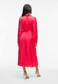 Abito in satin rosso con vita cinturata, maniche lunghe e design a lunghezza media. Tessuto liscio e leggermente trasparente, con polsini con bottoni.