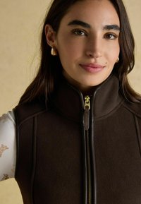 Braunfarbener Fleece-Weste mit hohem Kragen, goldener Reißverschluss und Lederbesatz. Getragen über einem leicht gemusterten Langarmhemd.