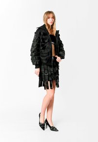 Chaqueta bomber negra con textura similar a cinta, falda negra con flecos a juego y tacones puntiagudos y brillantes; diseño estructurado y estilizado.