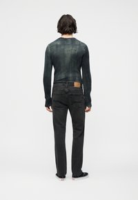 Άτομο που στέκ εται γυρισμένο προς τα πίσω, φοράει μακρυμάνικο, ξεθωριασμένο πράσινο πουλόβερ, μαύρα τζιν με πίσω τσέπες και μαύρα χαμηλά αθλητικά παπούτσια.