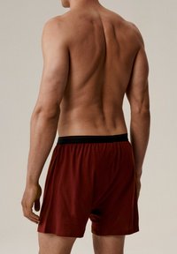 Czerwone majtki typu boxer z czarną elastyczną talią, prezentowane na nagich plecach, ukazujące gładką fakturę i luźny krój.