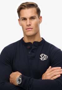 Marineblaues Langarm-Henley-Shirt mit geripptem Kragen, drei Knöpfen und gesticktem Logo. Das Modell trägt eine schwarze Uhr mit Lederband.